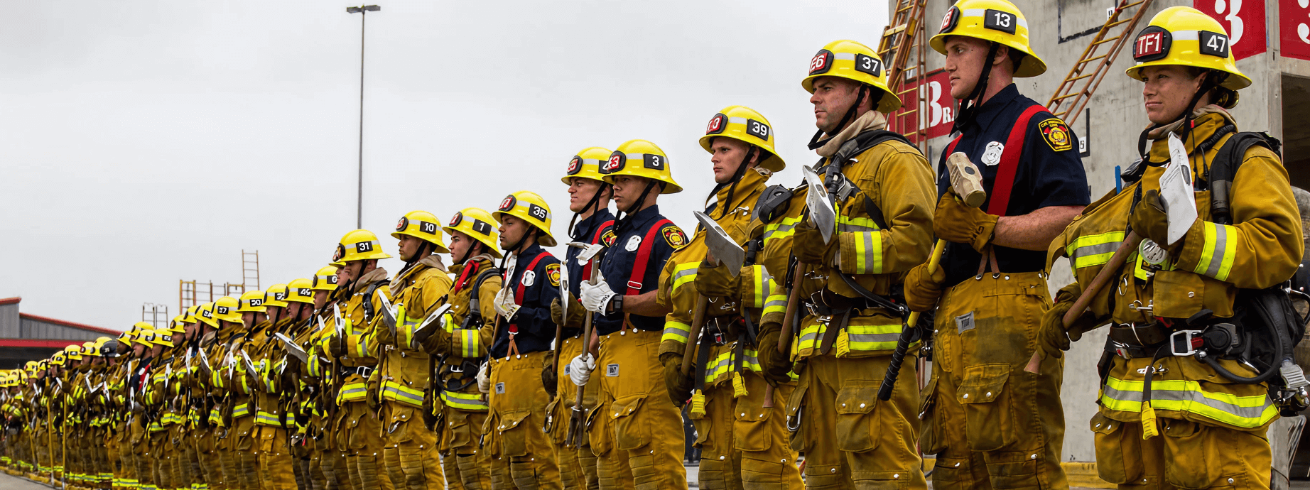 A group of uniformed firefighters stands in a formation, dressed in full turnout gear including yellow helmets and protective clothing. Many are holding firefighting tools such as axes and Halligan bars. The setting appears to be an outdoor training facility with ladders and building props in the background, under a cloudy sky.